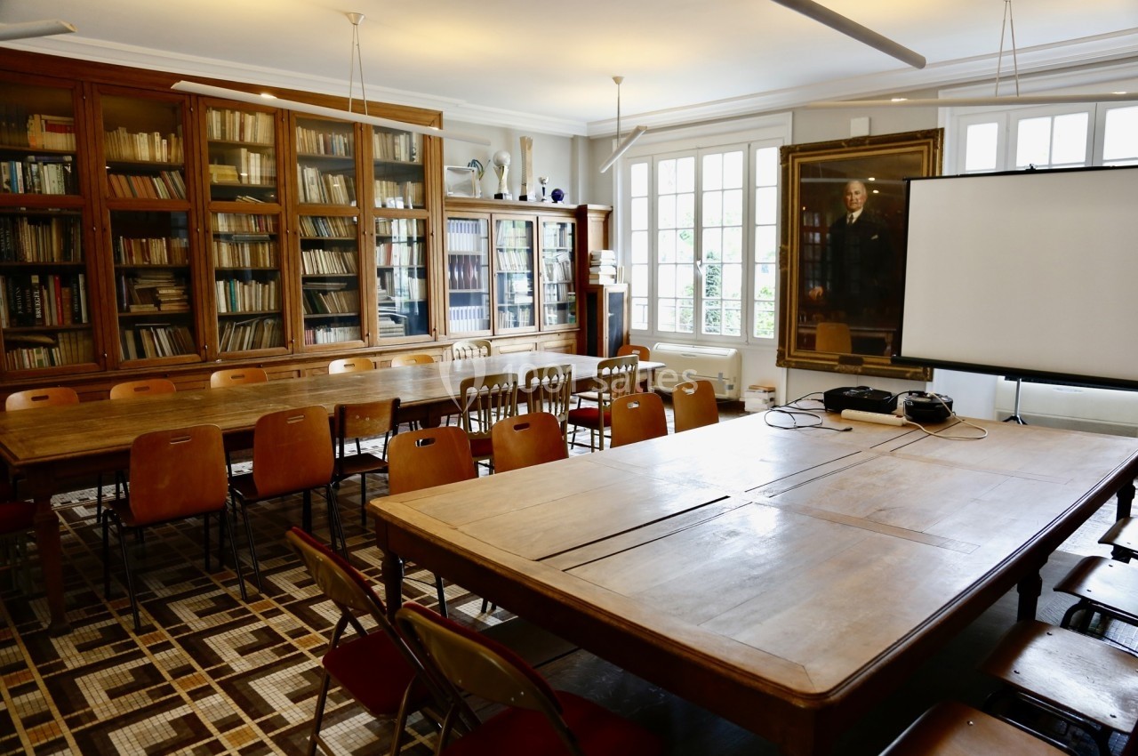 Salle de réunion avec tables en bois, chaises, bibliothèque remplie de livres et un écran de projection.