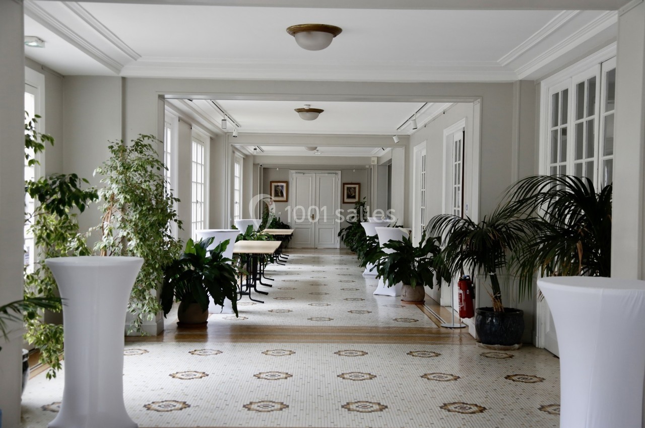 Couloir lumineux avec sol en mosaïque, plantes vertes, tables et chaises, encadré de grandes fenêtres.