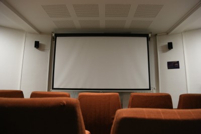 Écran de projection blanc dans une petite salle équipée de fauteuils marron et de haut-parleurs muraux.