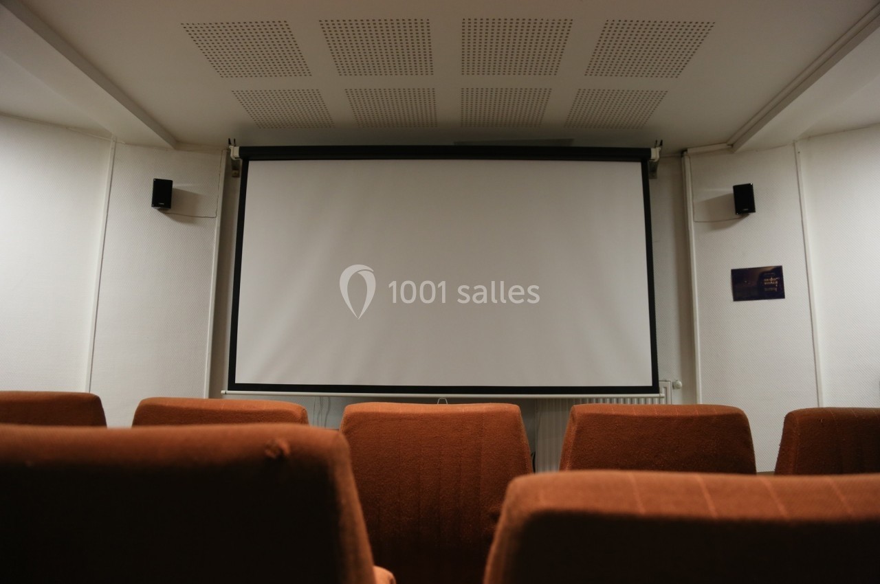 Écran de projection blanc dans une petite salle équipée de fauteuils marron et de haut-parleurs muraux.