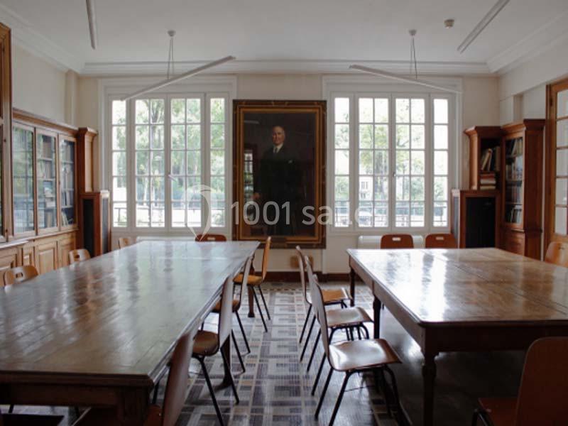 Salle lumineuse avec grandes fenêtres, tables en bois, chaises et un portrait accroché au mur central.