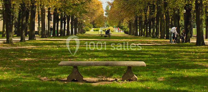 Banc en pierre au premier plan dans une allée bordée d'arbres, avec des promeneurs visibles au loin.