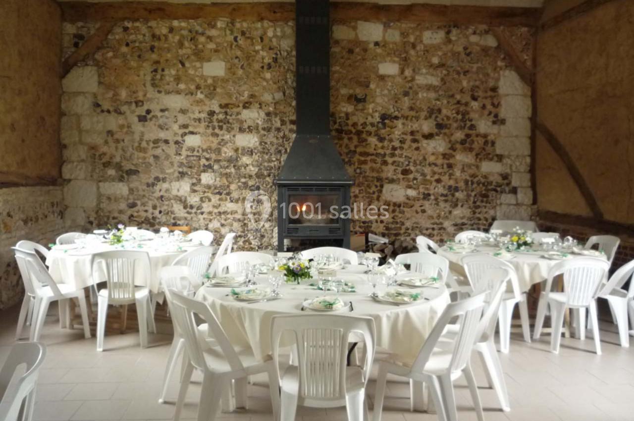 Location salle Montivilliers (Seine-Maritime) - Ferme d'Epaville #9 Salle rustique avec murs en pierre, cheminée allumée et tables rondes dressées pour un repas, entourées de chaises blanches.
