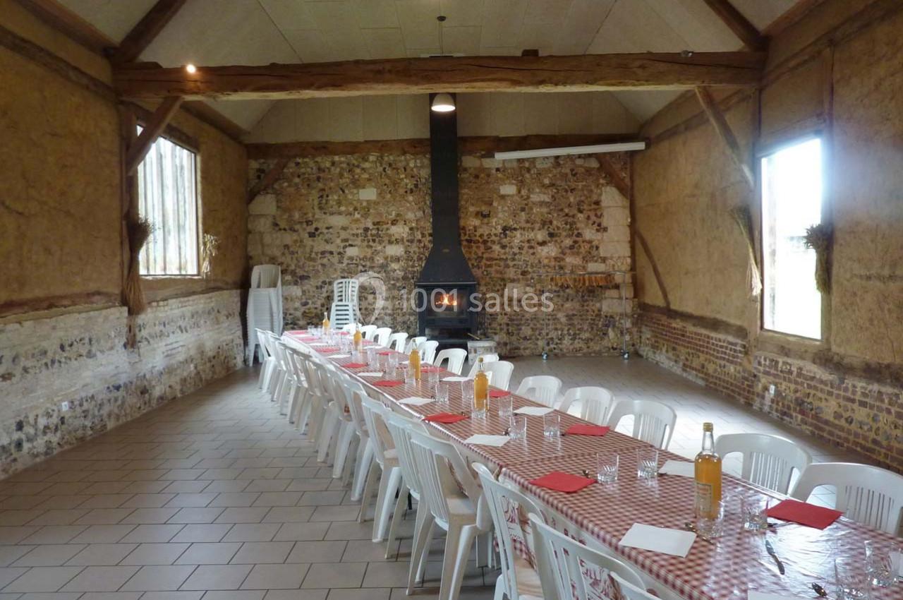 Location salle Montivilliers (Seine-Maritime) - Ferme d'Epaville #19 Grande salle rustique avec murs en pierre, cheminée allumée et longues tables dressées avec nappes à carreaux rouges.