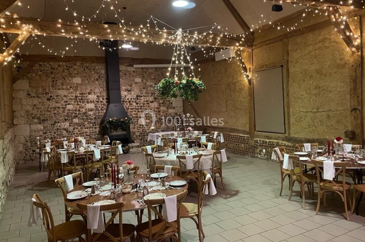 Location salle Montivilliers (Seine-Maritime) - Ferme d'Epaville #3 Salle de réception rustique avec tables rondes dressées, guirlandes lumineuses suspendues et murs en pierre apparente.