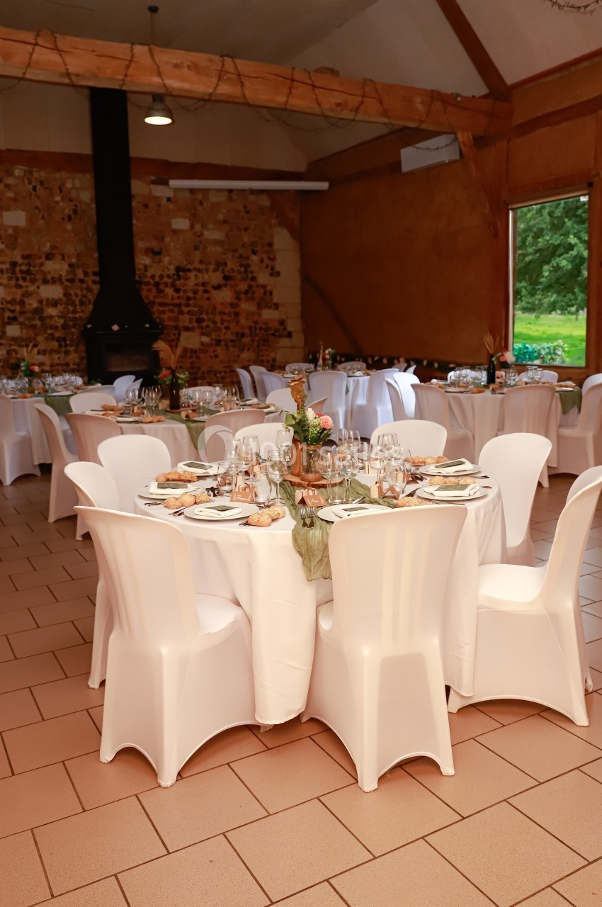 Location salle Montivilliers (Seine-Maritime) - Ferme d'Epaville #36 Salle de réception avec tables rondes dressées, nappes blanches, chaises assorties et décoration florale.