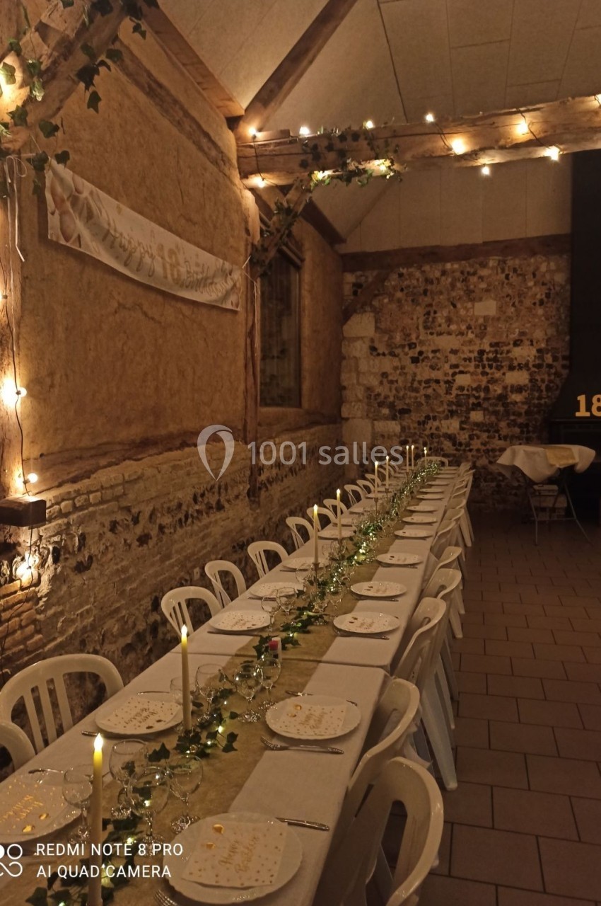 Location salle Montivilliers (Seine-Maritime) - Ferme d'Epaville #42 Grande table décorée pour un dîner, avec bougies et guirlandes lumineuses, dans une salle en pierres apparentes.