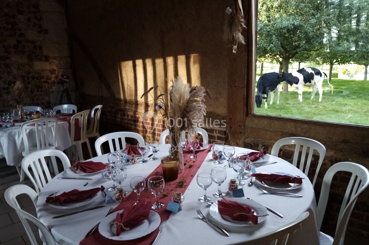 Location salle Montivilliers (Seine-Maritime) - Ferme d'Epaville #39 Salle de réception rustique avec table dressée et vue sur des vaches dans un pré à travers une grande fenêtre.