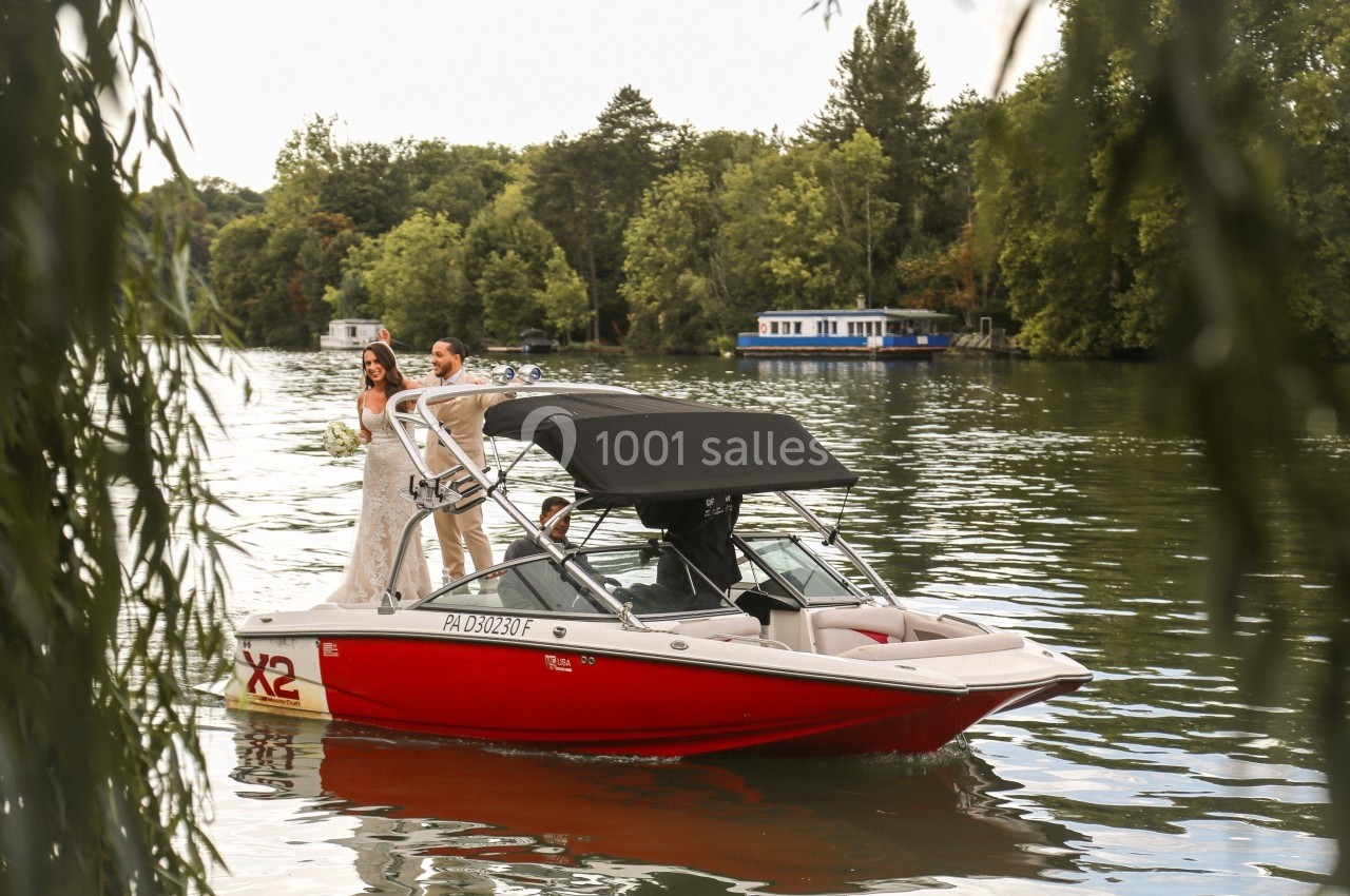 Un couple debout sur un bateau rouge naviguant sur une rivière entourée de végétation et de péniches.
