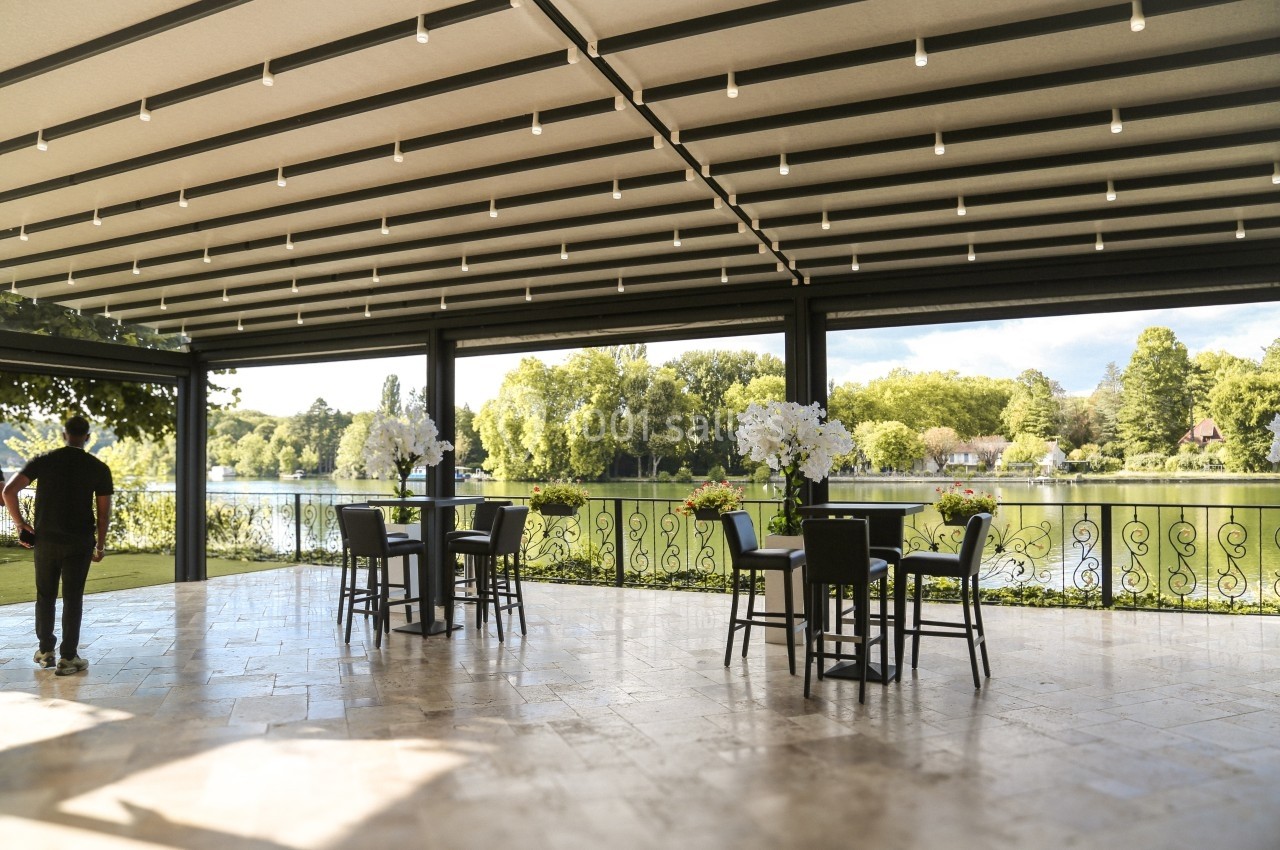 Terrasse couverte avec vue sur un lac et un parc, aménagée avec des tables hautes et des chaises noires.