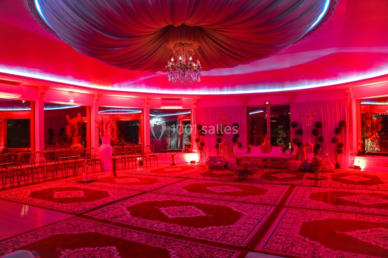 Salle de réception décorée de tapis rouges, éclairée par des lumières roses et un lustre central.