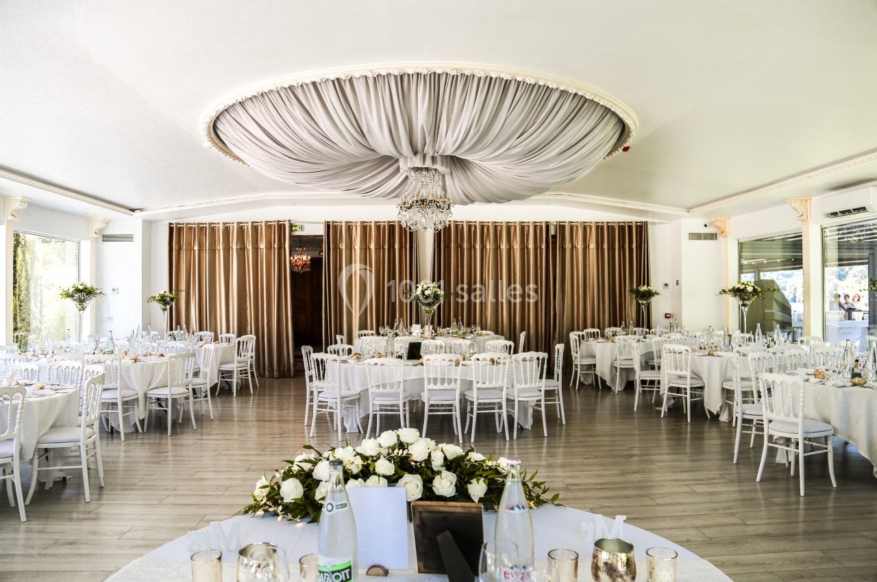 Salle de réception lumineuse avec tables rondes dressées, chaises blanches et un grand lustre central sous un plafond drapé.