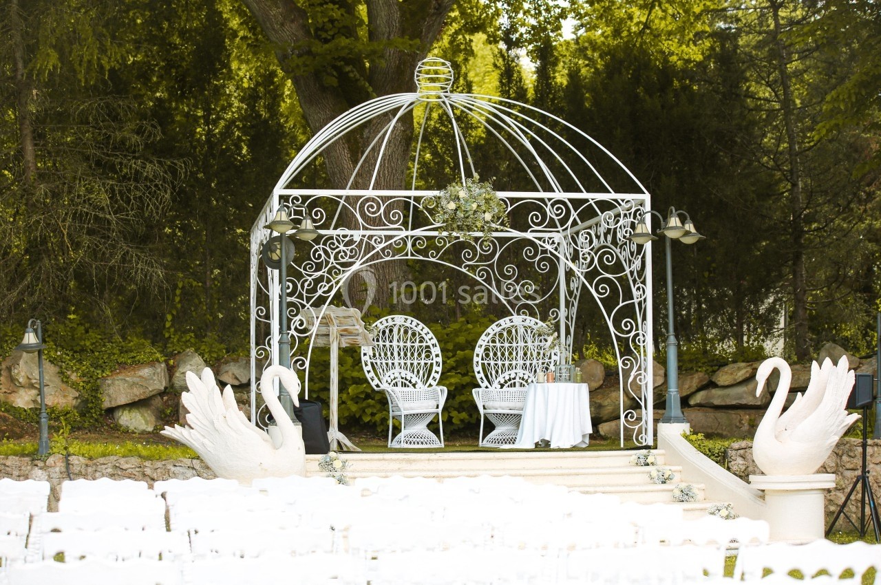 Kiosque en fer forgé décoré, avec deux chaises blanches et une table, entouré de statues de cygnes et d'arbres.