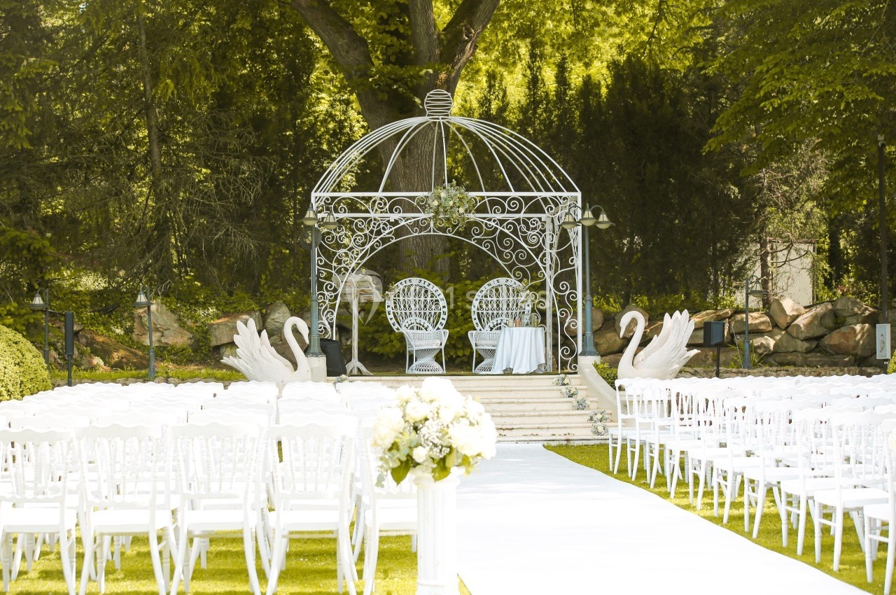 Allée bordée de chaises blanches menant à une arche métallique ornée, entourée de verdure et décorée de cygnes.
