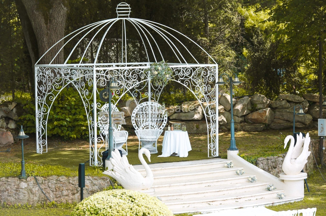 Kiosque en fer forgé blanc avec chaises et table, entouré de verdure et décoré de sculptures de cygnes.