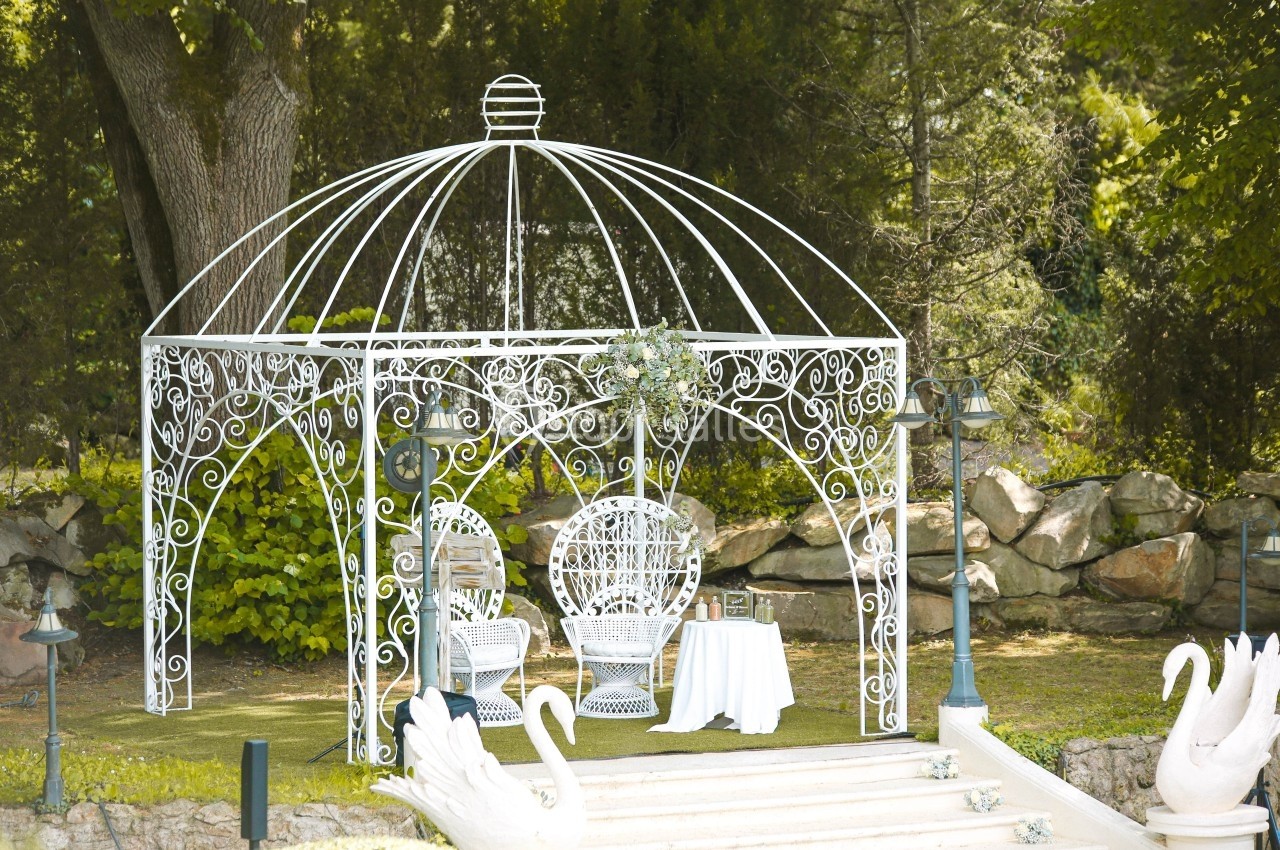 Tonnelle en fer forgé blanc avec deux fauteuils assortis, table décorée et cygnes en premier plan, dans un jardin verdoyant.