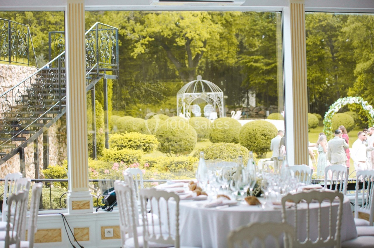 Vue sur un jardin avec des arbustes taillés, une tonnelle blanche et des invités, depuis une salle de réception élégante.