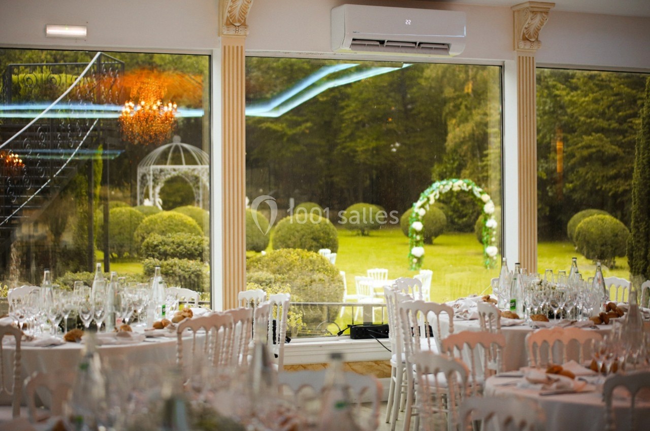 Location salle Le Coudray-Montceaux (Essonne) - Le Manoir des Cygnes #12 Salle de réception élégante avec tables dressées, vue sur un jardin verdoyant et une arche décorée pour une cérémonie.