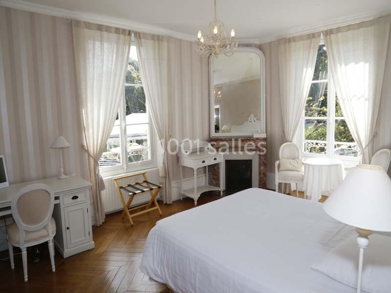 Chambre lumineuse avec lit double, bureau, cheminée, grandes fenêtres et vue sur un jardin.