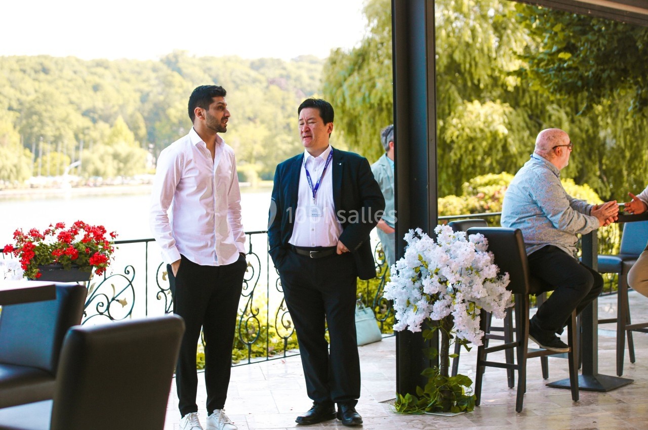 Deux hommes discutent debout sur une terrasse avec vue sur un lac, entourés de fleurs et de mobilier de jardin.