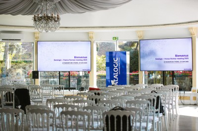 Salle lumineuse avec des stands promotionnels, des affiches et des tables recouvertes de nappes bleues, entourées de chaises…