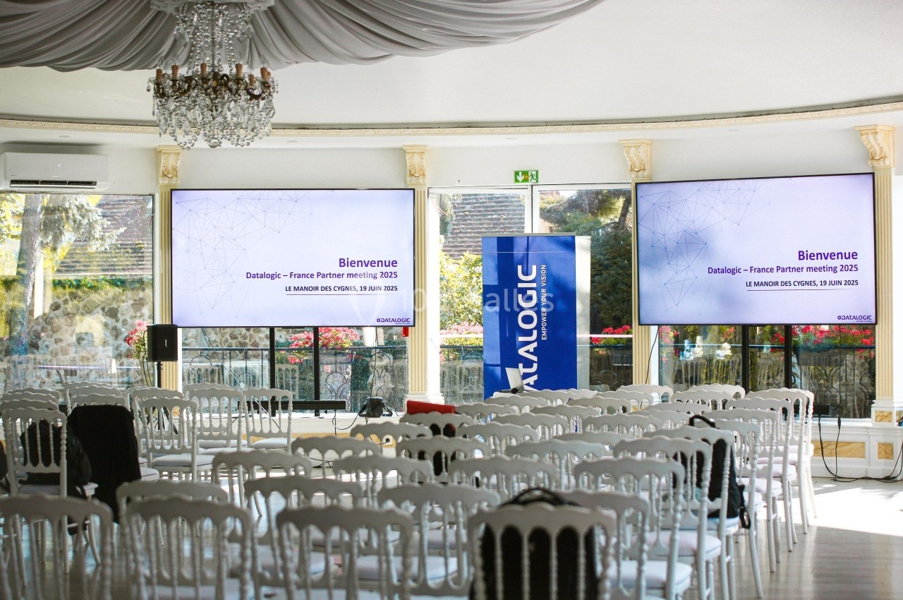 Salle de conférence lumineuse avec des chaises blanches alignées, deux écrans affichant un message de bienvenue.