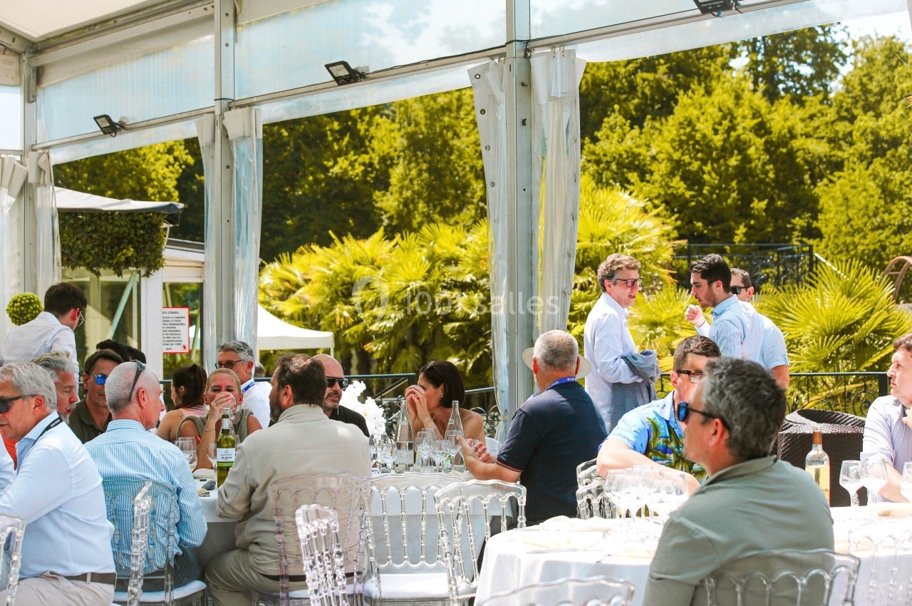 Des personnes assises et debout discutent autour de tables dressées sous une tente, avec un jardin en arrière-plan.