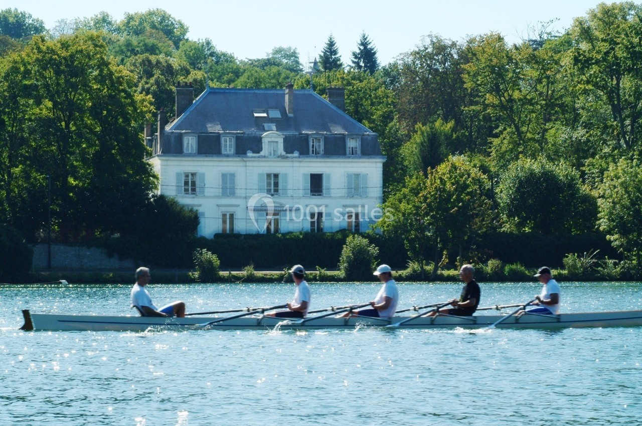 Location salle Le Coudray-Montceaux (Essonne) - Le Manoir des Cygnes #16 Quatre personnes rament sur une rivière devant une grande maison blanche entourée d'arbres.