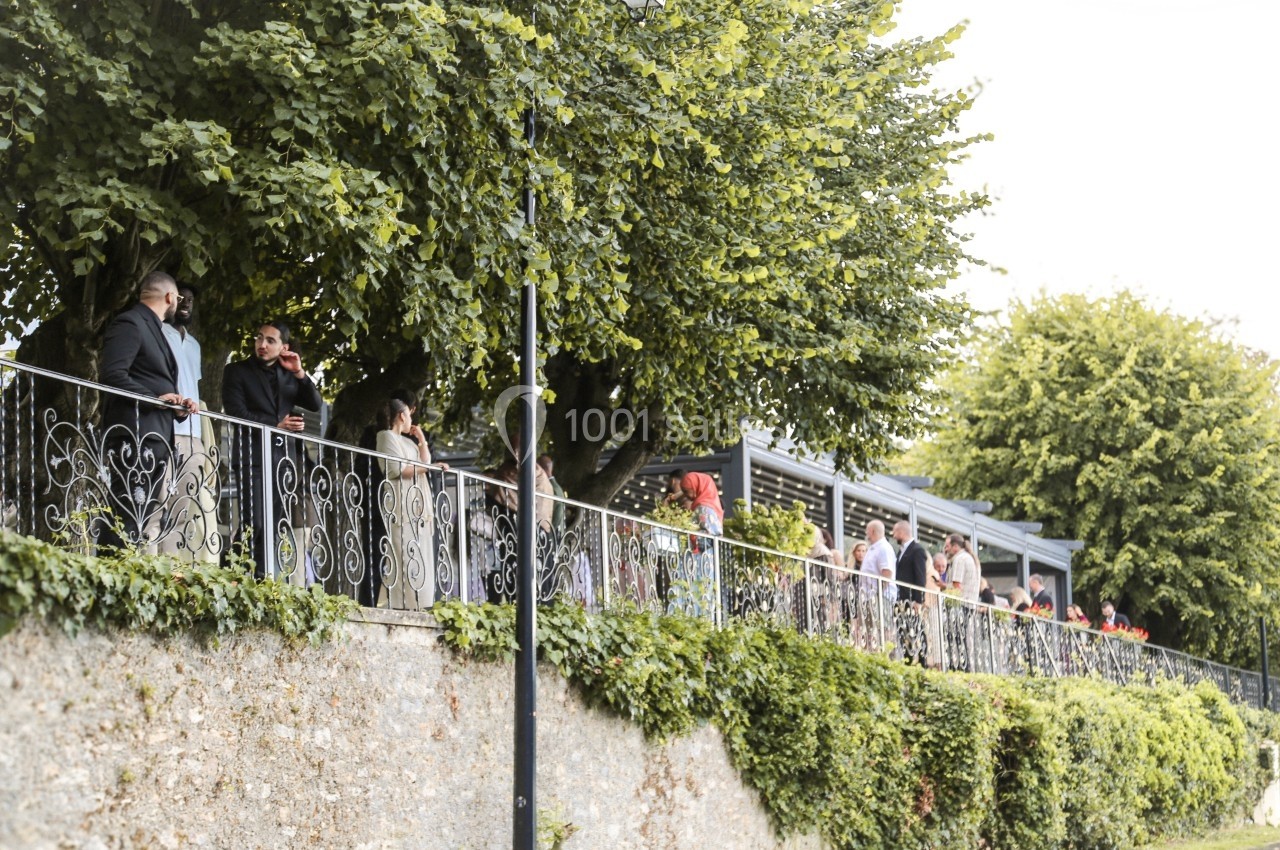 Location salle Le Coudray-Montceaux (Essonne) - Le Manoir des Cygnes #13 Des personnes discutent et se promènent le long d'une balustrade entourée de verdure et d'arbres.