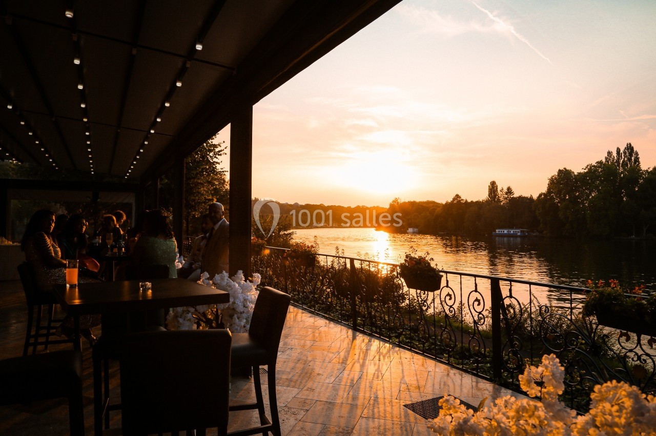 Location salle Le Coudray-Montceaux (Essonne) - Le Manoir des Cygnes #6 Terrasse au bord d'une rivière au coucher du soleil, avec des tables, des chaises et des fleurs en premier plan.
