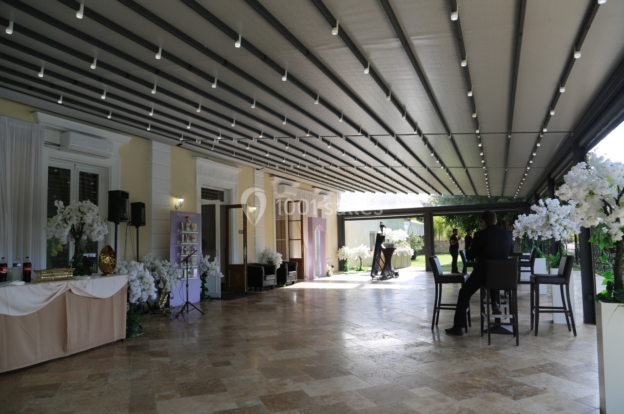 Location salle Le Coudray-Montceaux (Essonne) - Le Manoir des Cygnes #11 Terrasse couverte avec éclairage suspendu, tables, chaises et décorations florales, donnant sur un jardin verdoyant.