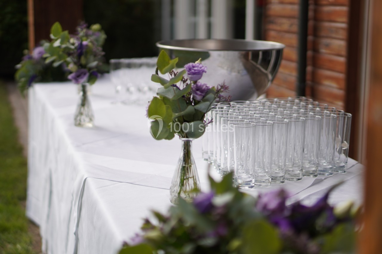 Table dressée à l'extérieur avec des verres alignés, des vases de fleurs violettes et un grand saladier en métal.