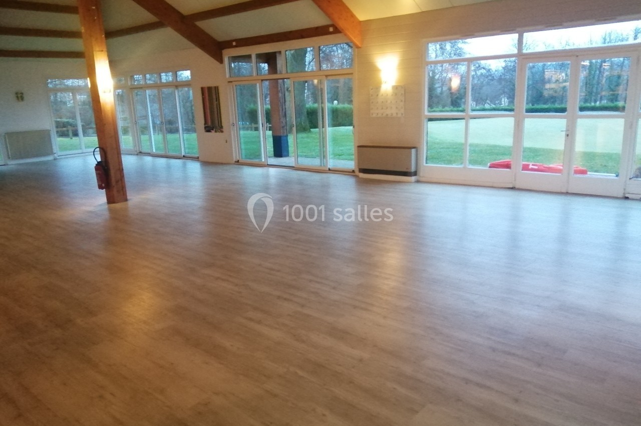 Salle vide avec parquet clair, grandes baies vitrées donnant sur un espace extérieur verdoyant.