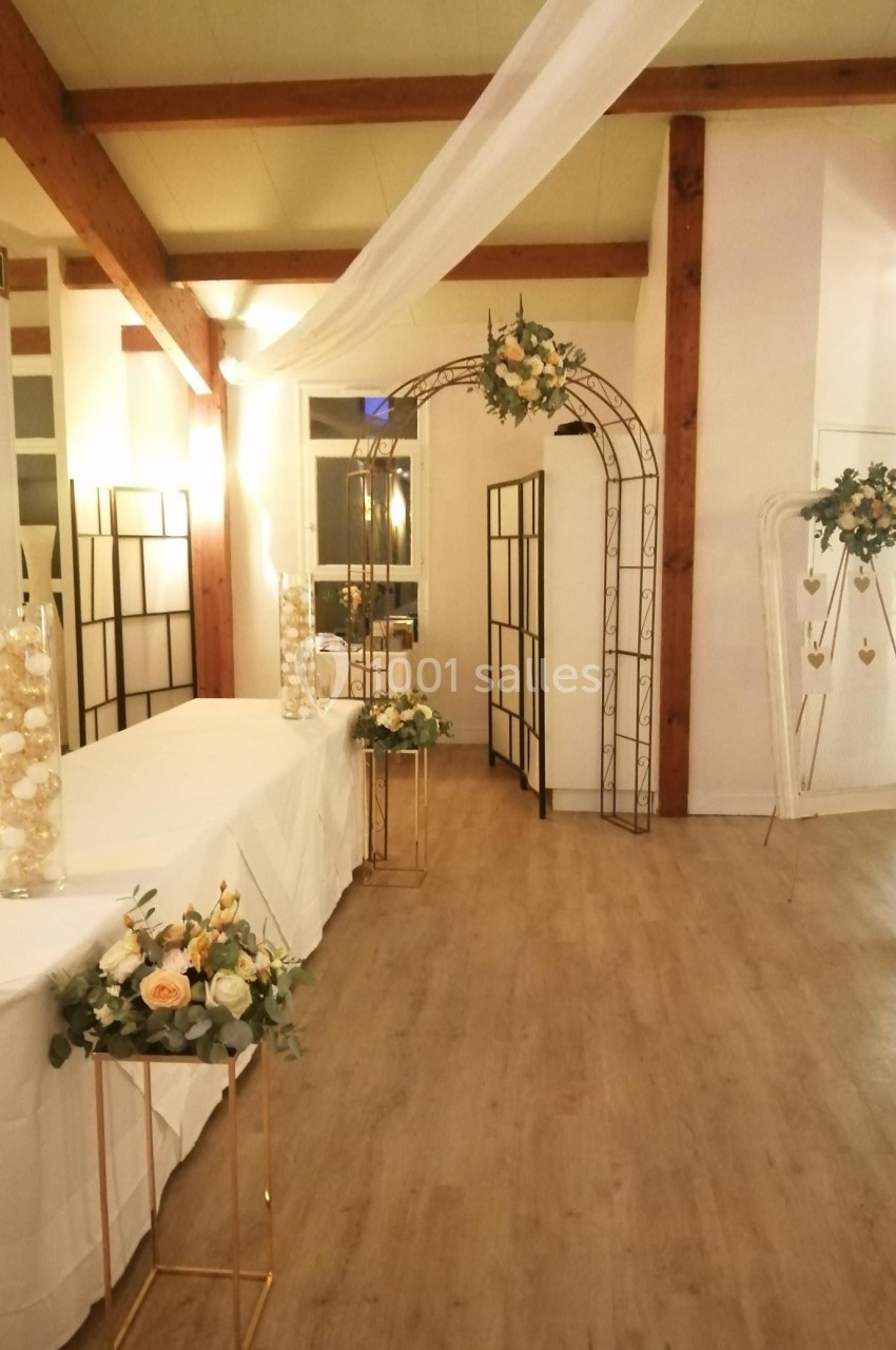 Salle décorée pour un événement avec arche florale, tables blanches et arrangements de fleurs.