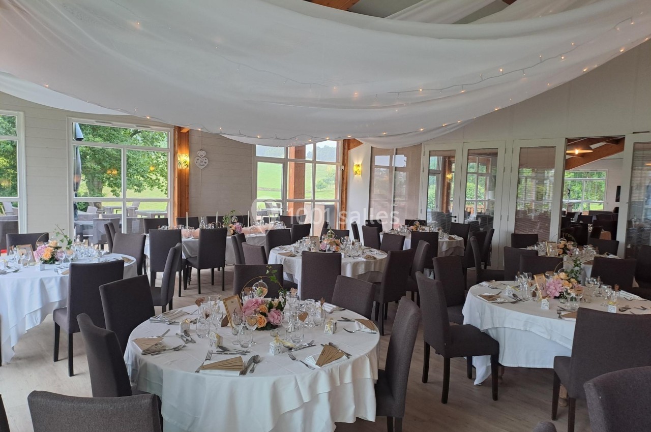 Salle de réception décorée avec des tables rondes dressées, nappes blanches, fleurs et guirlandes lumineuses au plafond.
