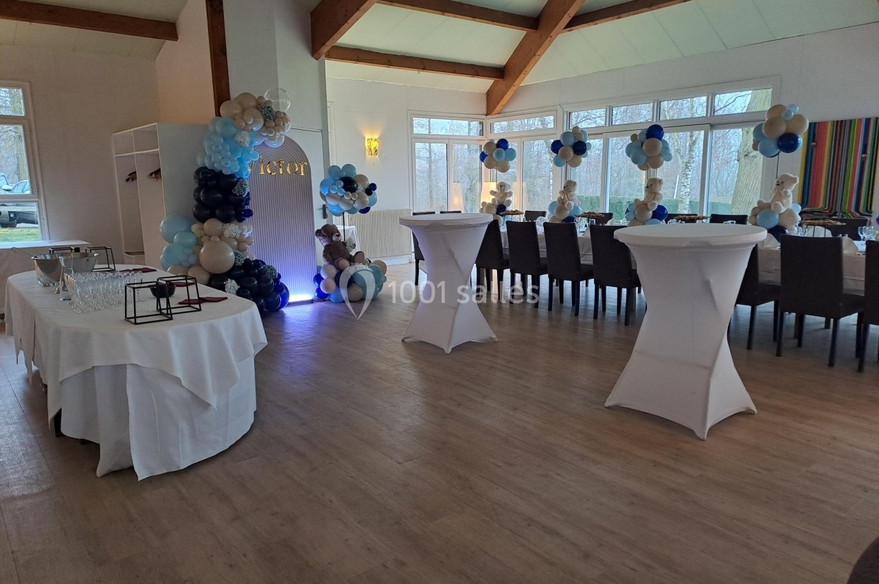 Salle décorée pour un événement avec tables dressées, ballons bleus et blancs, et grandes baies vitrées donnant sur l…