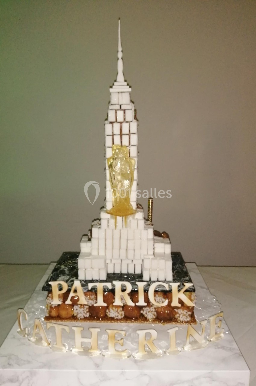 Gâteau en forme de gratte-ciel avec des noms ’Patrick’ et ’Catherine’ inscrits en lettres dorées à la base.