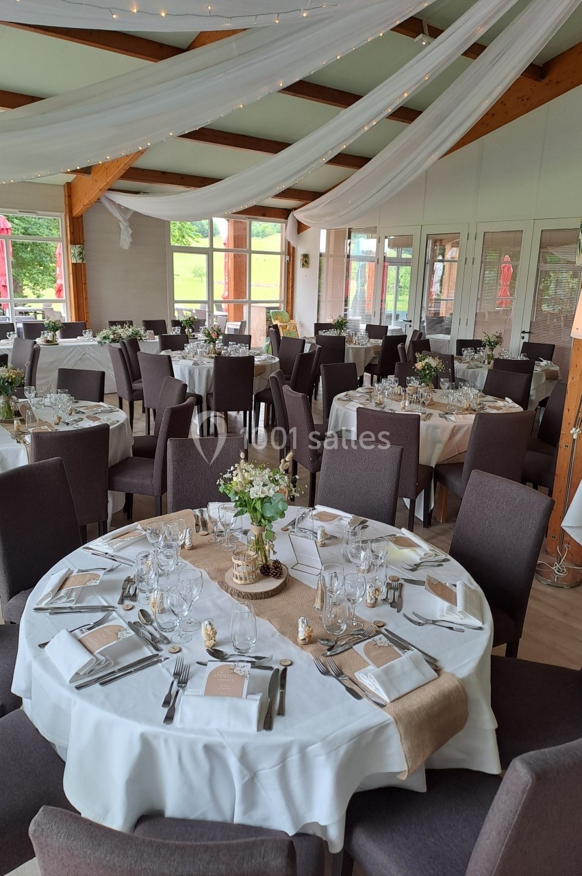 Salle de réception décorée avec des tables rondes dressées, nappes blanches, centres de table floraux et voilages suspendus.