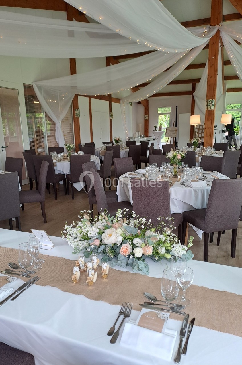 Salle de réception décorée pour un mariage avec tables dressées, nappes blanches, fleurs et voilages suspendus.