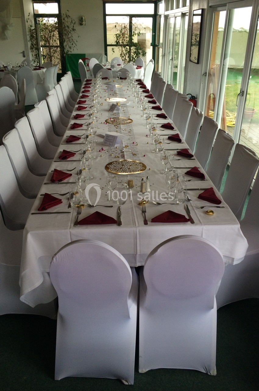 Grande table rectangulaire dressée pour un repas, avec nappes blanches, serviettes rouges et chaises couvertes.