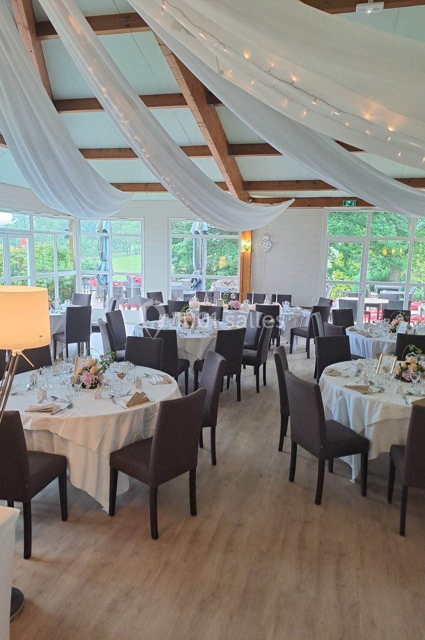 Salle de réception décorée avec des tables rondes dressées, chaises noires et voilages suspendus au plafond.