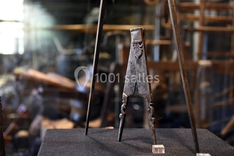 Compas ancien en métal exposé sur un support noir, avec un arrière-plan flou d'atelier ou de forge.