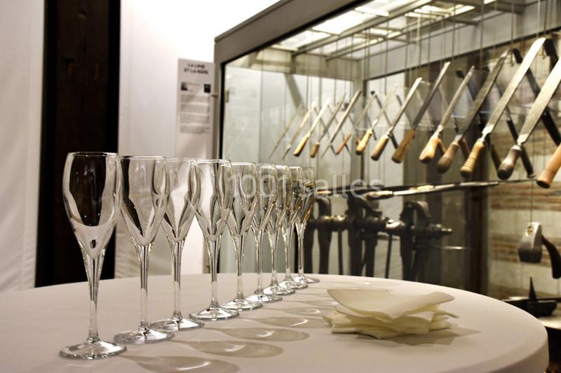 Rangée de flûtes à champagne sur une table ronde devant une vitrine exposant des outils anciens.