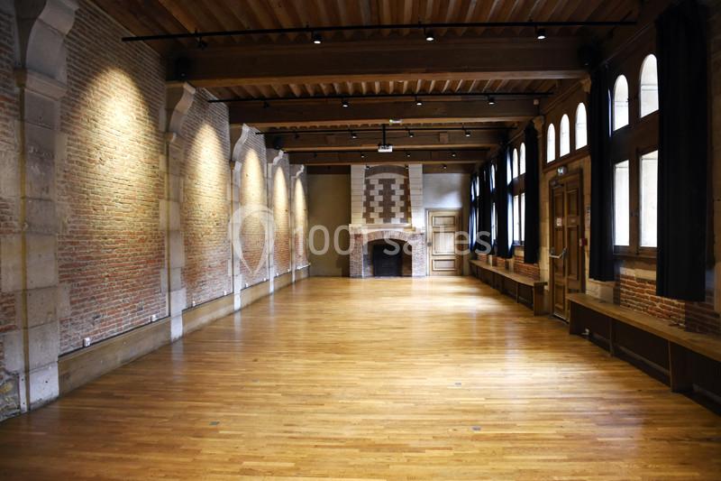 Salle vide avec murs en briques, parquet en bois, plafond en poutres apparentes et cheminée en pierre au fond.