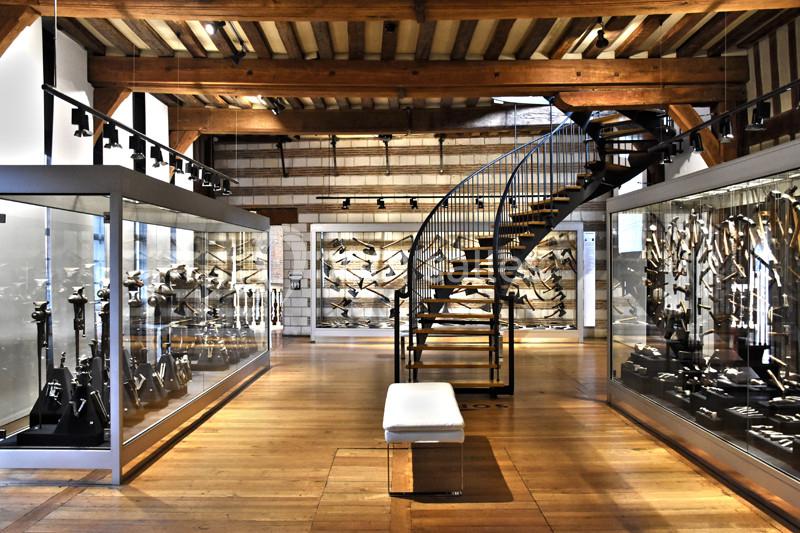 Salle d'exposition avec vitrines contenant des armes anciennes, escalier en colimaçon et plafond en poutres apparentes.