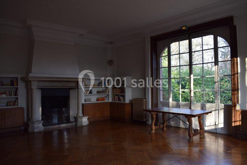 Salle lumineuse avec parquet en bois, cheminée, étagères remplies de livres et grande baie vitrée donnant sur un jardin.