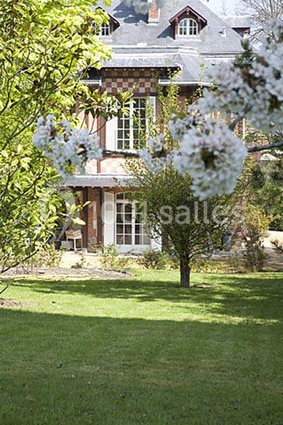 Maison en briques et colombages entourée d'un jardin verdoyant avec des arbres en fleurs au premier plan.