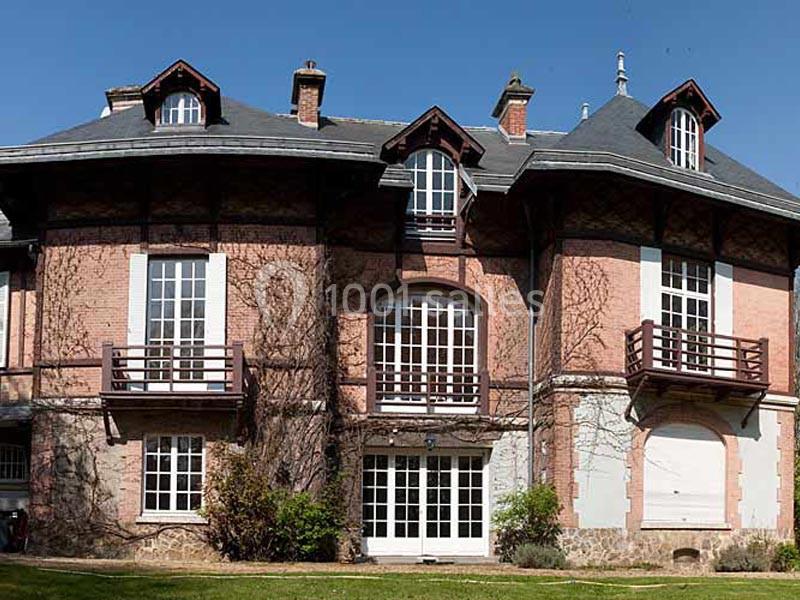 Façade d'une grande maison en briques avec des balcons en bois, des fenêtres cintrées et des plantes grimpantes.