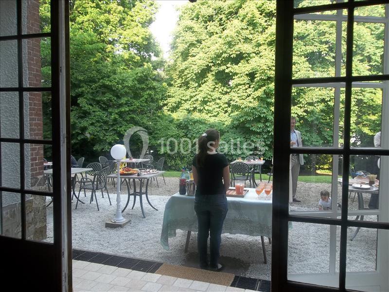Une femme debout près d'une table dressée, vue depuis une porte ouverte donnant sur une terrasse et un jardin verdoyant.