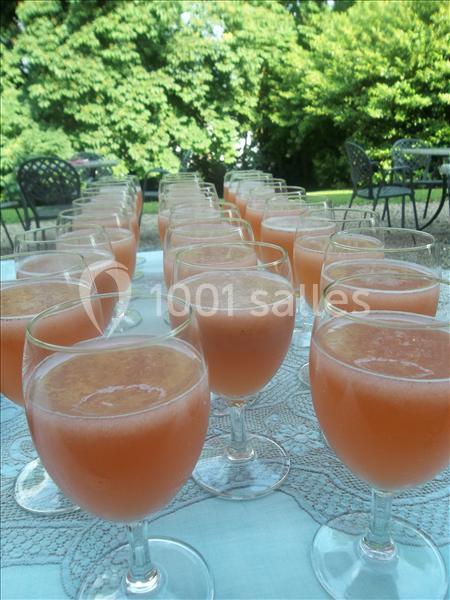 Verres de jus de fruits rose disposés sur une table en extérieur, avec des arbres et des chaises en arrière-plan.